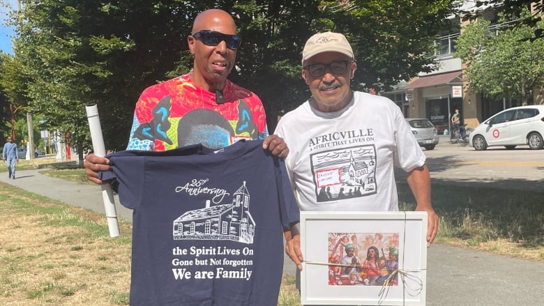 Two men are smiling while posing for the camera. They're both holding up gifts that they exchanged with each other. 