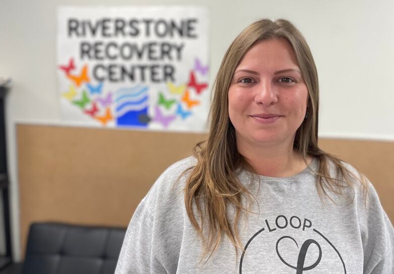 A woman with long blond hair wearing a grey crewneck sweater. A posterboard on the wall behind her says 