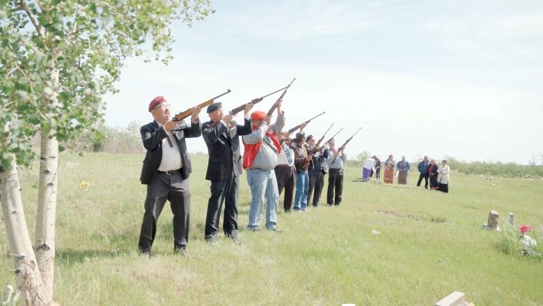 A line up of men, some in military garb, shoot rifles in the air while standing outside near a cemetery. A small group of onlookers is in the distant background.