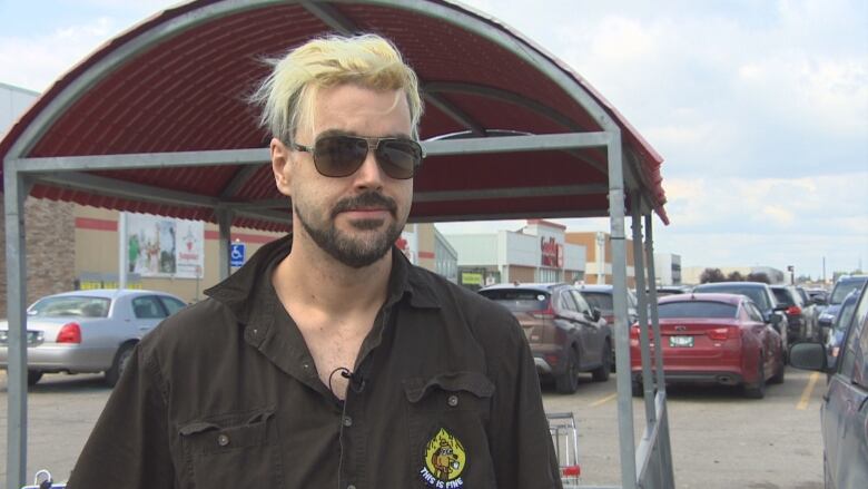 A man wearing sunglasses stands in a parking lot in front of a shopping cart corral.