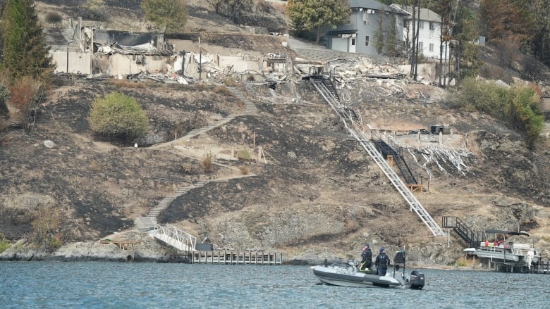 An RCMP boat is seen on a lake. In the foreground is a hilly area with debris and destroyed houses.