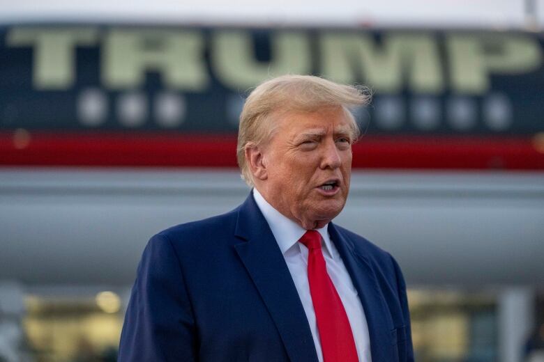 A man in a suit and tie is shown in closeup speaking while in front of a plane adorned with the name 'Trump.'