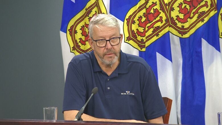 A man with white hair and a blue polo shirt wears glasses. He sits in front of Nova Scotia flags. 