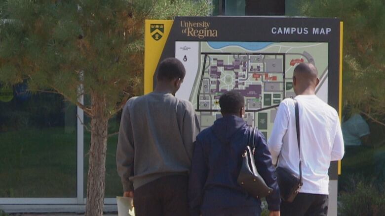 Three Black people stand, looking at a campus map of the University of Regina. They are outside and the sun is shining.