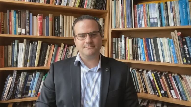 A man in a suit and open-collar shirt sits in front of shelves heavy with books.