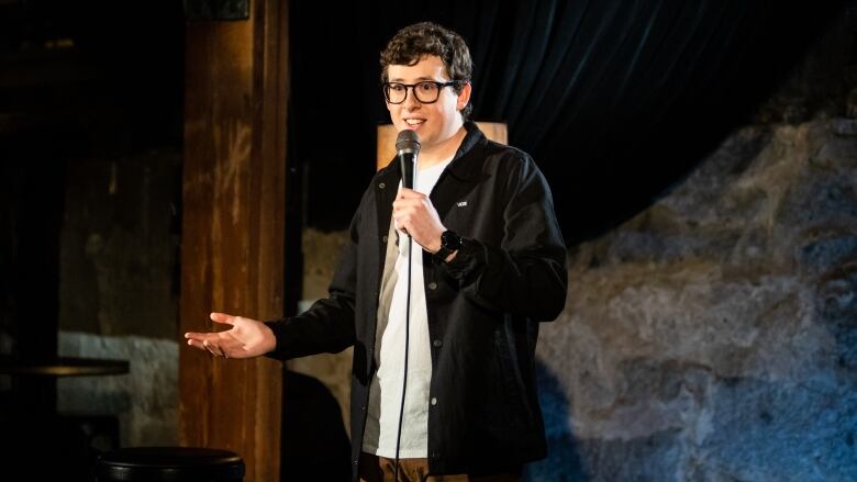 A person with short hair and glasses speaks into a microphone on a stage.