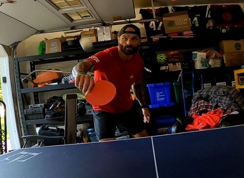 A bearded man in red shirt play table tennis in a garage.