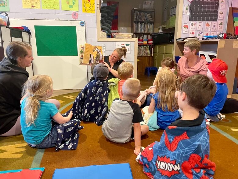 A group of small children sit in a group of from of their teacher who is reading them a book. 