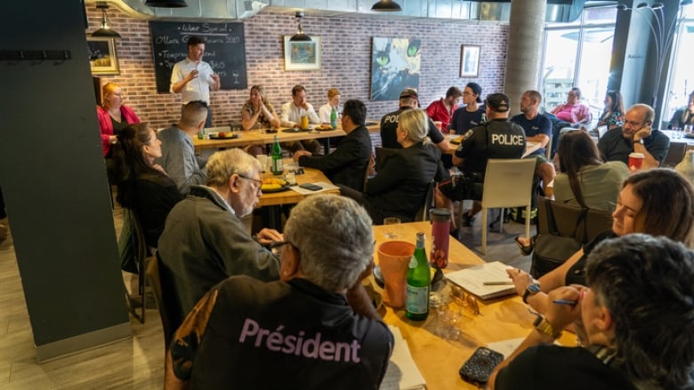 People sit inside a restaurant. A man in a white shirt stands as he speaks to the crowd. 