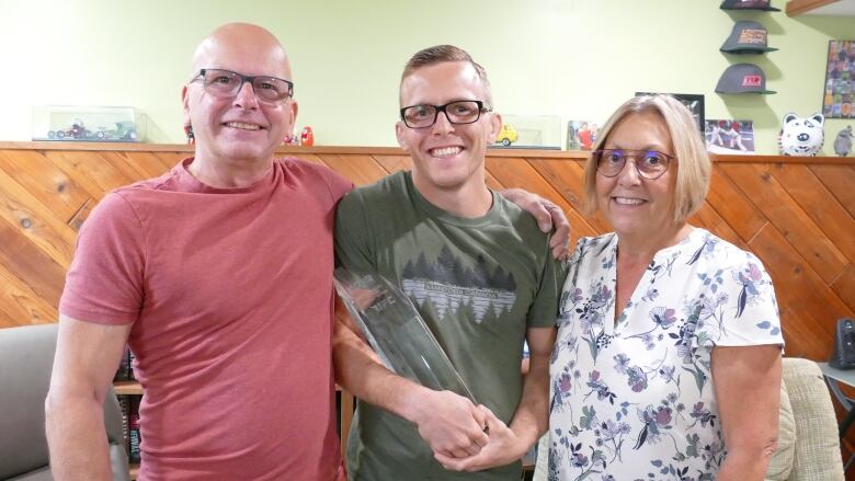 Two men and a woman pose for a photo. The younger man, in the middle, holds a glass trophy.