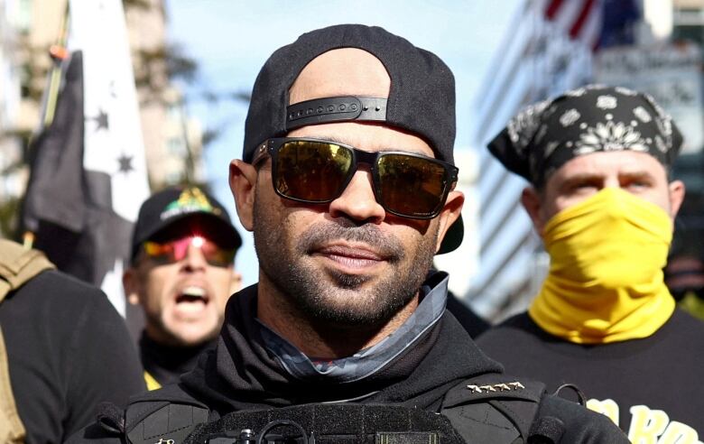 A man is seen in closeup outdoors wearing sunglasses and a baseball cap turned backwards.