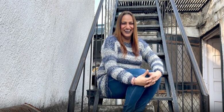 Still frame from the film I Am Sirat. Sirat Taneja grins widely, sitting on a fire escape.