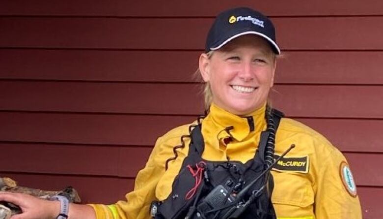 A white woman with a black ball cap and a yellow high-vis jacket stands beside a wood pile