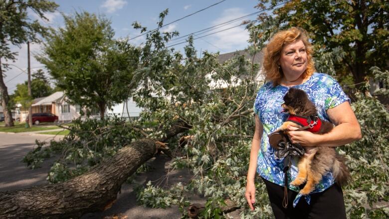 A woman holding a small dog stands in front of a downed tree