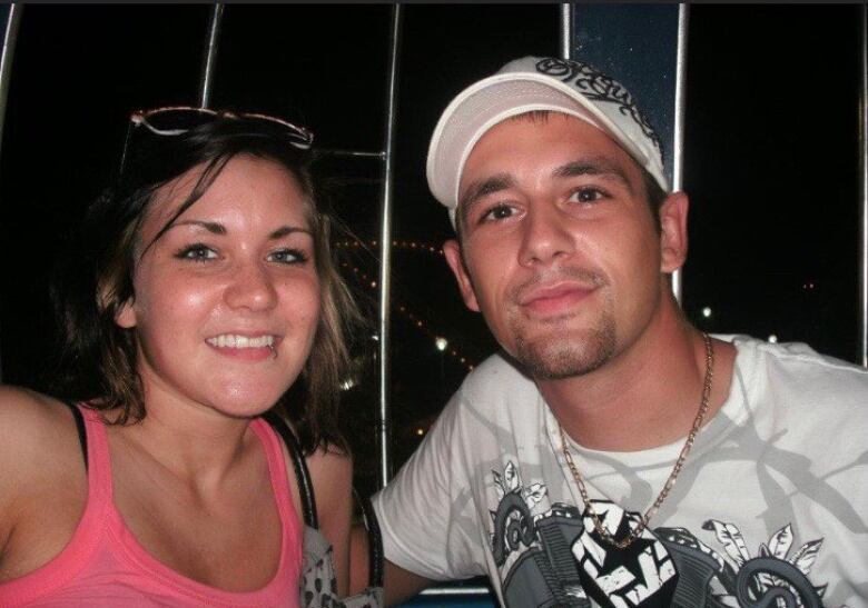 A man and a woman smile. The man is wearing a white shirt and baseball hat. 