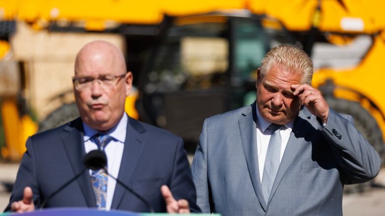 A man in a suit speaks at a lectern while another, standing to one side, puts his hand to his forehead.  