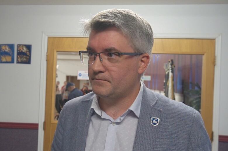 An unsmiling man with glasses, wearing a grey dress shirt and grey blazer.