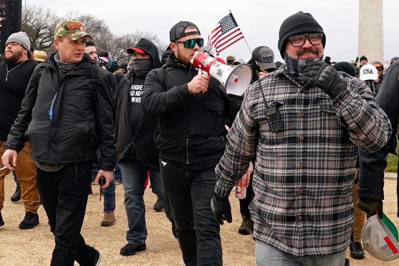 Three men in coats and various caps on their heads walk outdoors. The man in the middle appears to be speaking through a bullhorn.