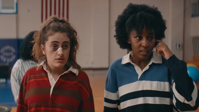 Two high school girls, bruised and bandaged, stand together in a gym.