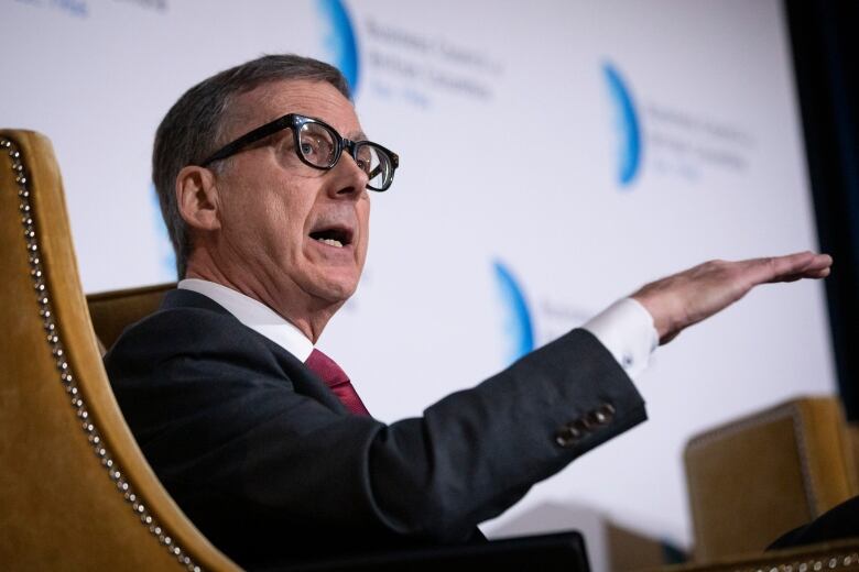 A man in suit speaks while raising his hand and sitting on a chair.