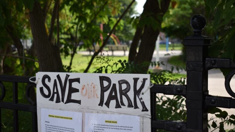 A 'Save Our Park' sign hangs on a fence near a park in summer.