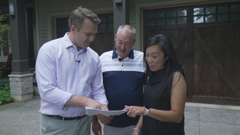 A man in a white button-down shirt holds a paper for a middle-aged man and woman to examine as they stand in the driveway of a home. 