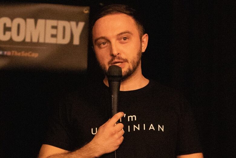 A man with dark hair and a neatly groomed beard and moustache wears a black t-shirt with the phrase 'I'm Ukrainian' as he speaks into a microphone on a stage. 