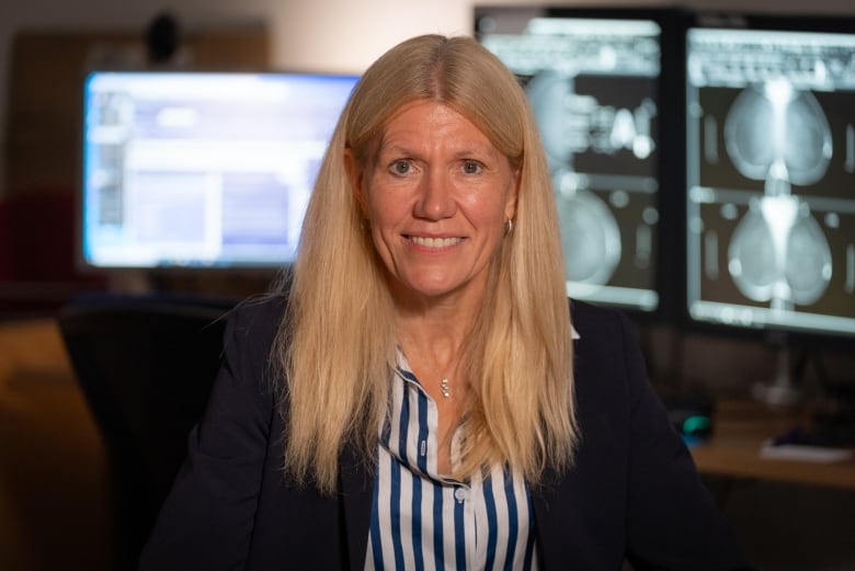 A woman sits smiling with screens on in the background showing breast imaging.