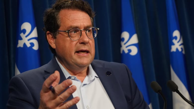 A man wearing glasses and a navy blue suit speaks at a podium in front of a Quebec flag. 