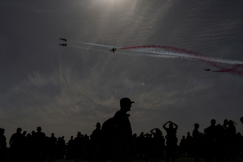 Spectators in silhouette watch aircraft performing in the sky, with colourful contrails. 