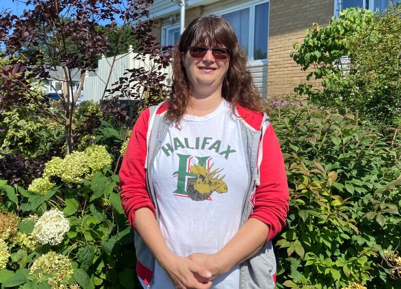 A woman stands in her garden. She's wearing a pink sweater, sunglasses and a Halifax Mooseheads shirt. Her house is behind her and her garden.