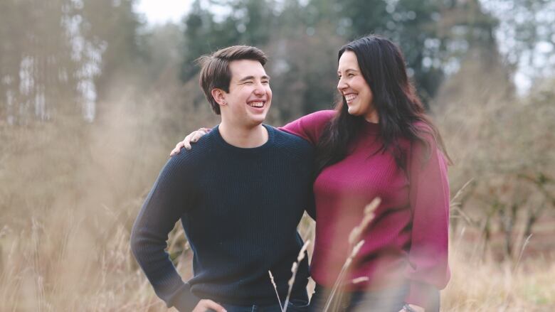 A young man and a woman stand arm-in-arm in a field. 