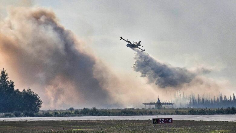 A plane drops water above a runway as a large cloud of smoke rises a short distance away.