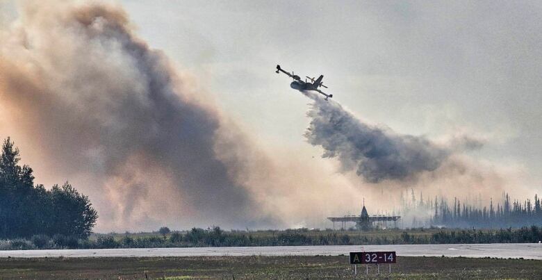 A plane drops water above a runway as a large cloud of smoke rises a short distance away.