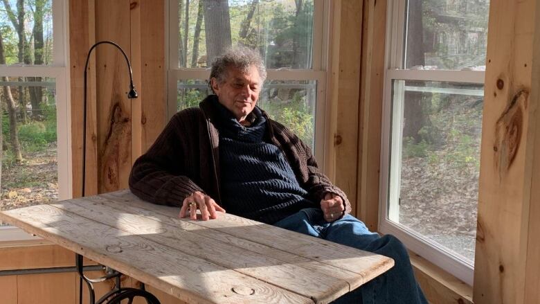 A man in his 70s smiles as he sits at a heritage wooden table in a wood cabin in a forested area.