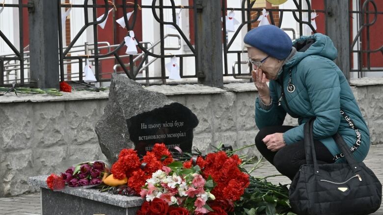 An older woman in a hat and coat crouches before a stone memorial with flowers in front of iron fencing.