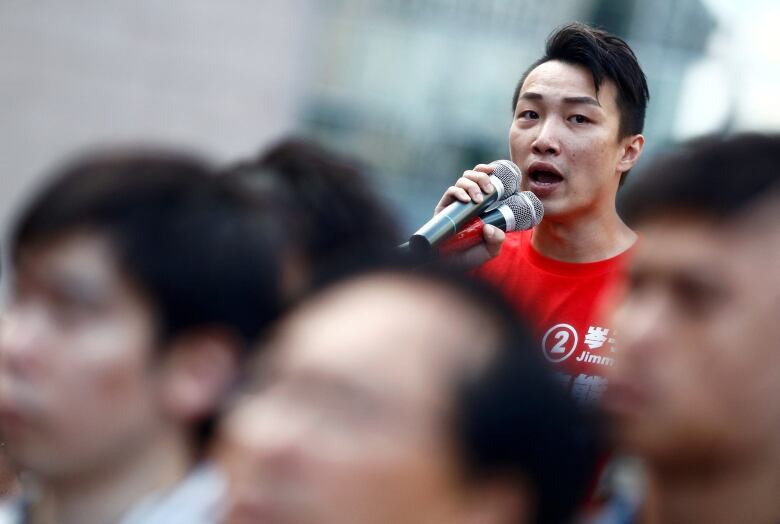 A man speaks into two microphones while standing in a crowd.