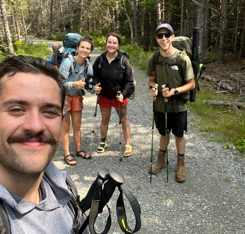 A group of people standing on a gravel hiking trail in hiking gear. A man on the left of the frame is close to the camera, holding it in selfie mode, while three others stand in a line further back.