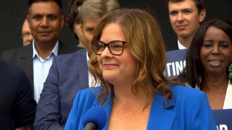 Woman with glasses wearing a blue suit speaks at a microphone with several people behind her. 