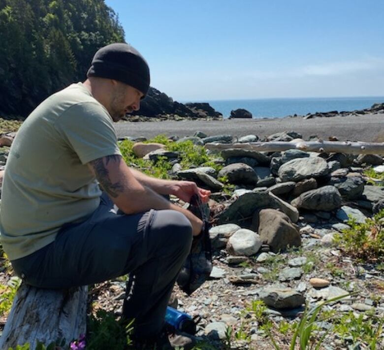 A man wearing a dark coloured tuque sits on a rock on the coast of the Bay of Fundy. He's wearing a light tee-shirt and dark coloured pants.