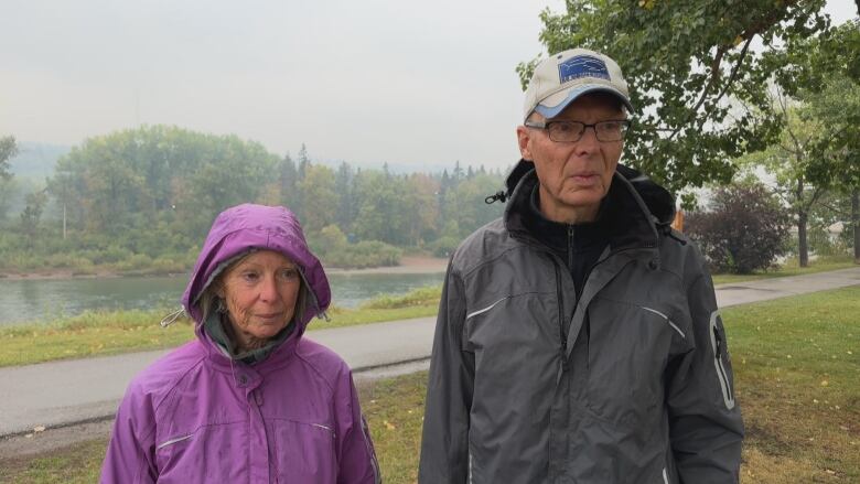 An older couple dressed in rain jackets stand in front of a river pathway during a smoky day in Calgary.