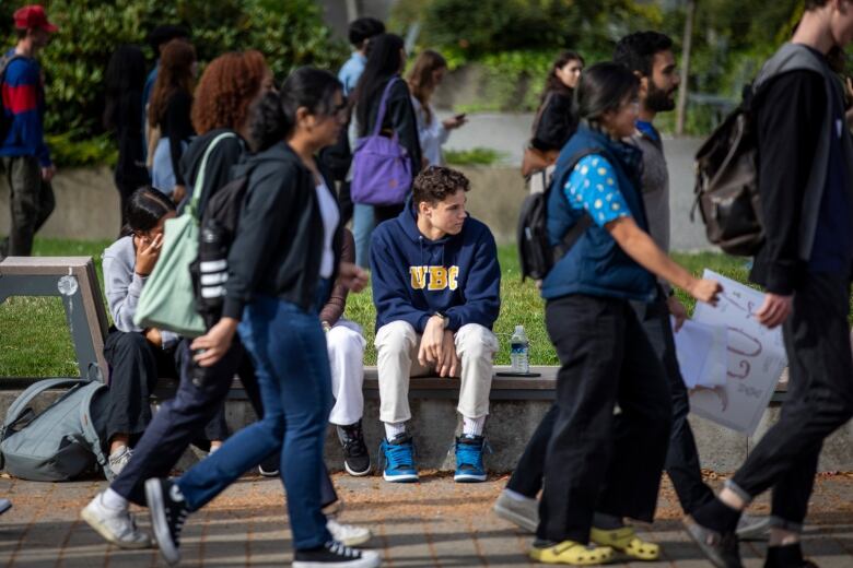 A crowd of students walk by and in the background is a student with a blue UBC hoodie sitting on a bench and looking to the side. 