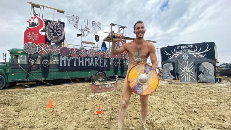 A naked man stands with a shield in front of himself holding a sword behind his back