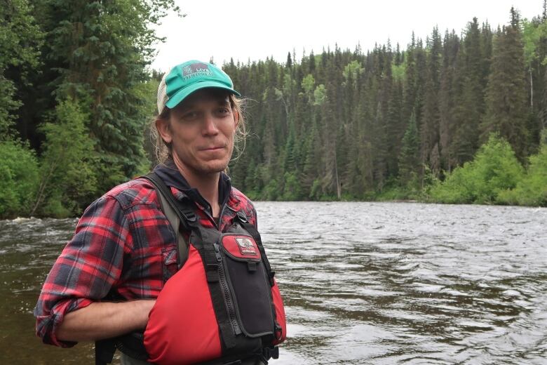 In a red and blue plade shirt a man stands in a river with his hands in a life jacket, smiling. 