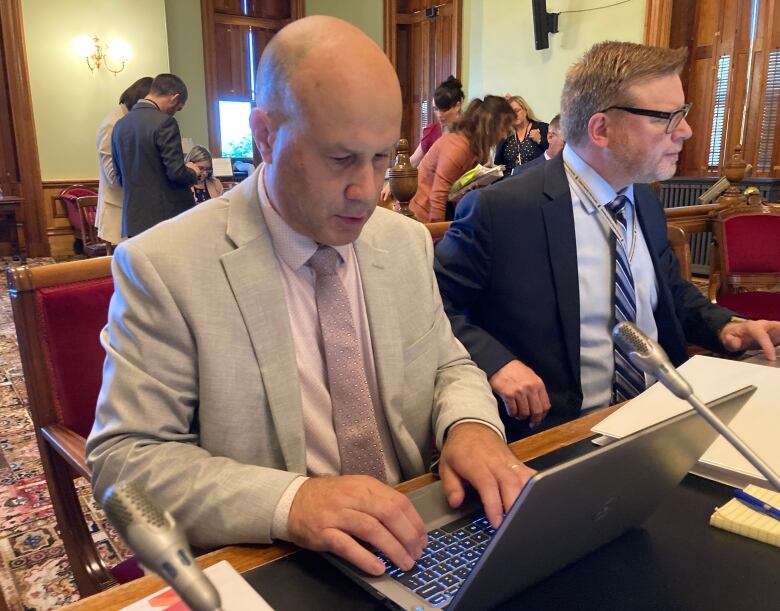 A man in a light grey suit and pink short types at a laptop on a desk in a committee room.