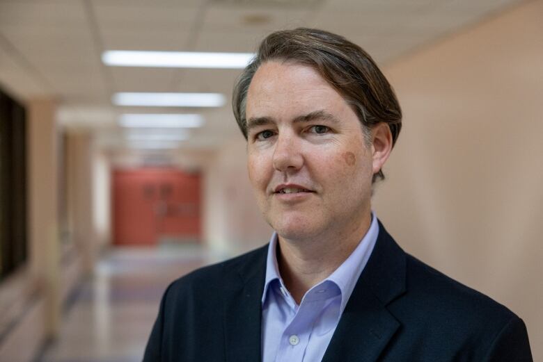 A male doctor in a blue shirt and navy suit is shown in the hallway of a hospital.