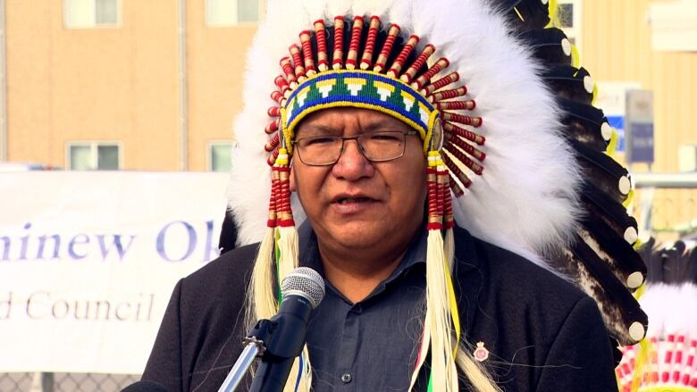 A First Nation chief wearing a traditional headdress speaks at a microphone. 