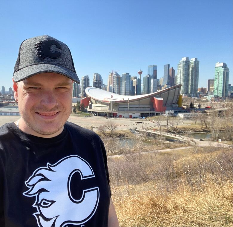 A man is seen smiling against the backdrop of downtown Calgary and the Scotiabank Saddledome.