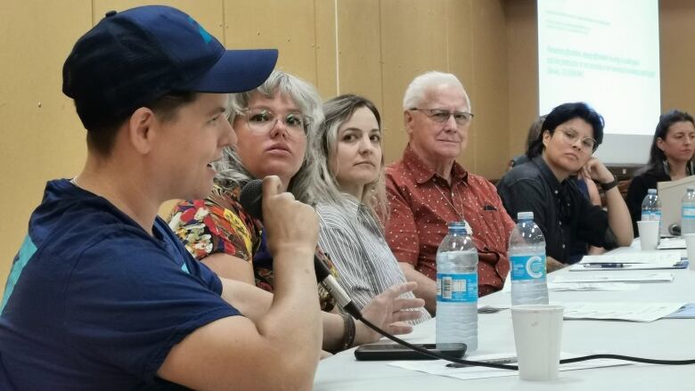 A number of people are sitting at a table. The person closest to the camera is holding a microphone and talking.