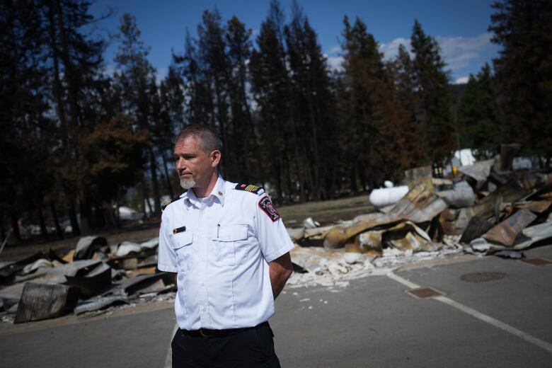 A white man wearing a white uniform stands in front of a burned-out building husk.